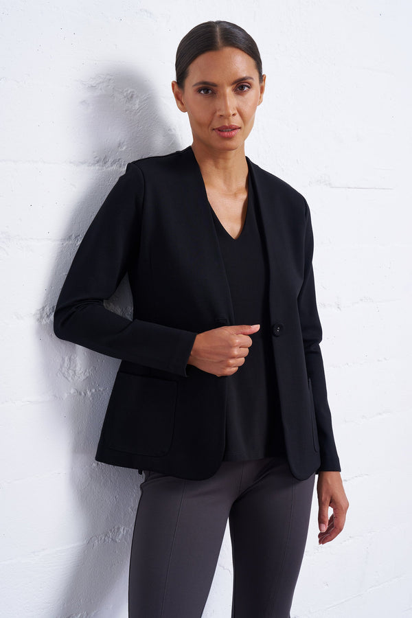 Woman wearing a black blazer and gray pants against a white wall